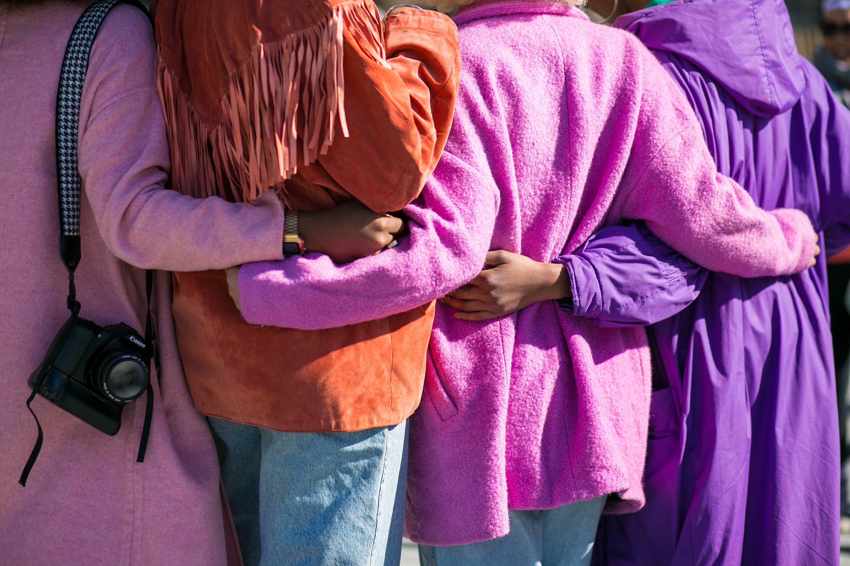 The backs of four people embracing one another in brightly-colored jackets, one of whom has a high quality camera around her shoulder